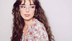 Model with clear frame glasses with dark curly hair.  Wearing a vintage flora blouse.