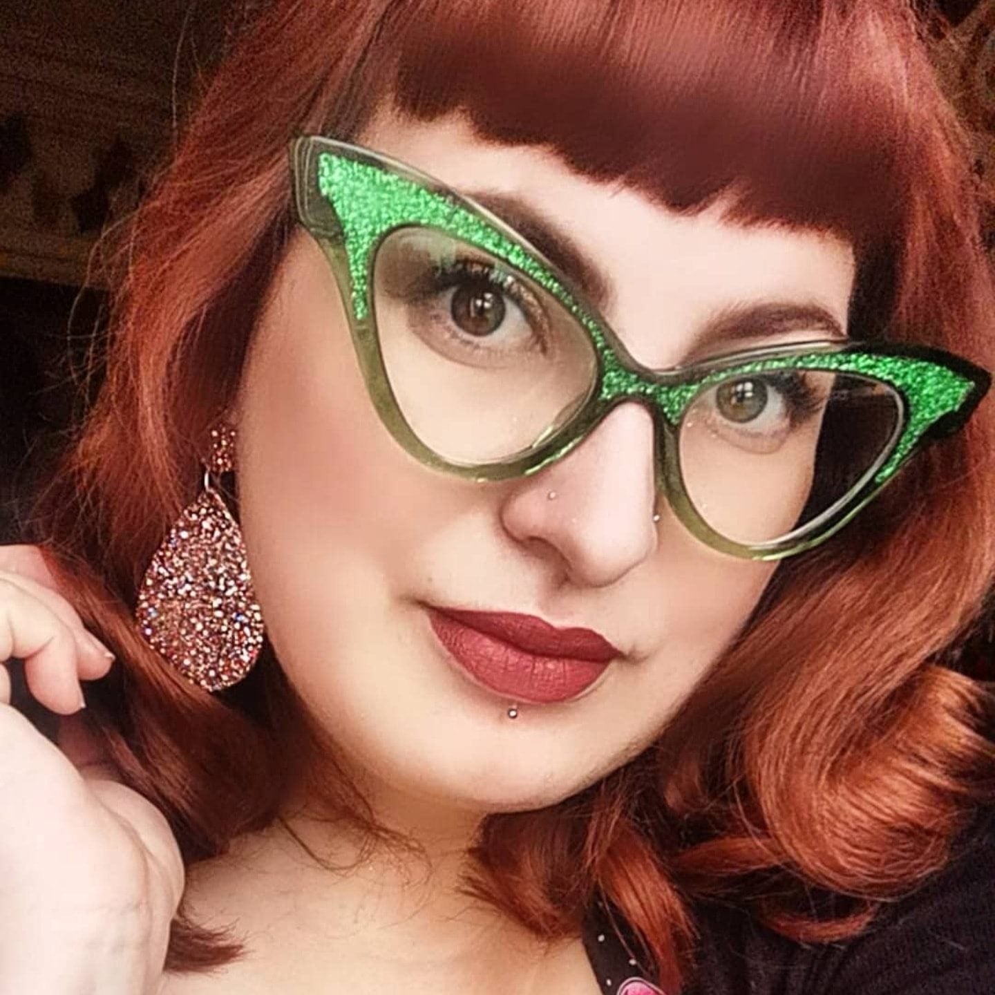 Person wearing green glittery cat-eye glasses with red hair and pink earrings.