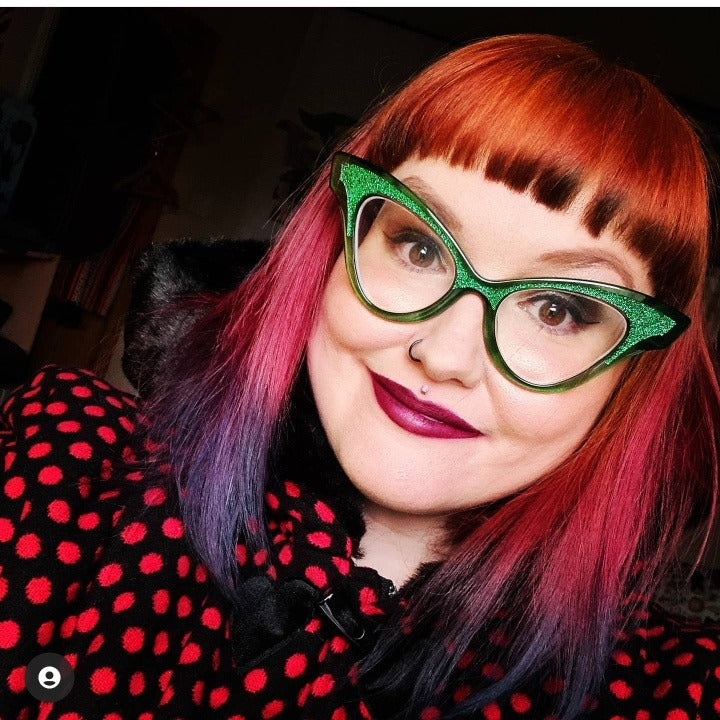 Person with red and black hair wearing green glasses and a polka dot outfit.