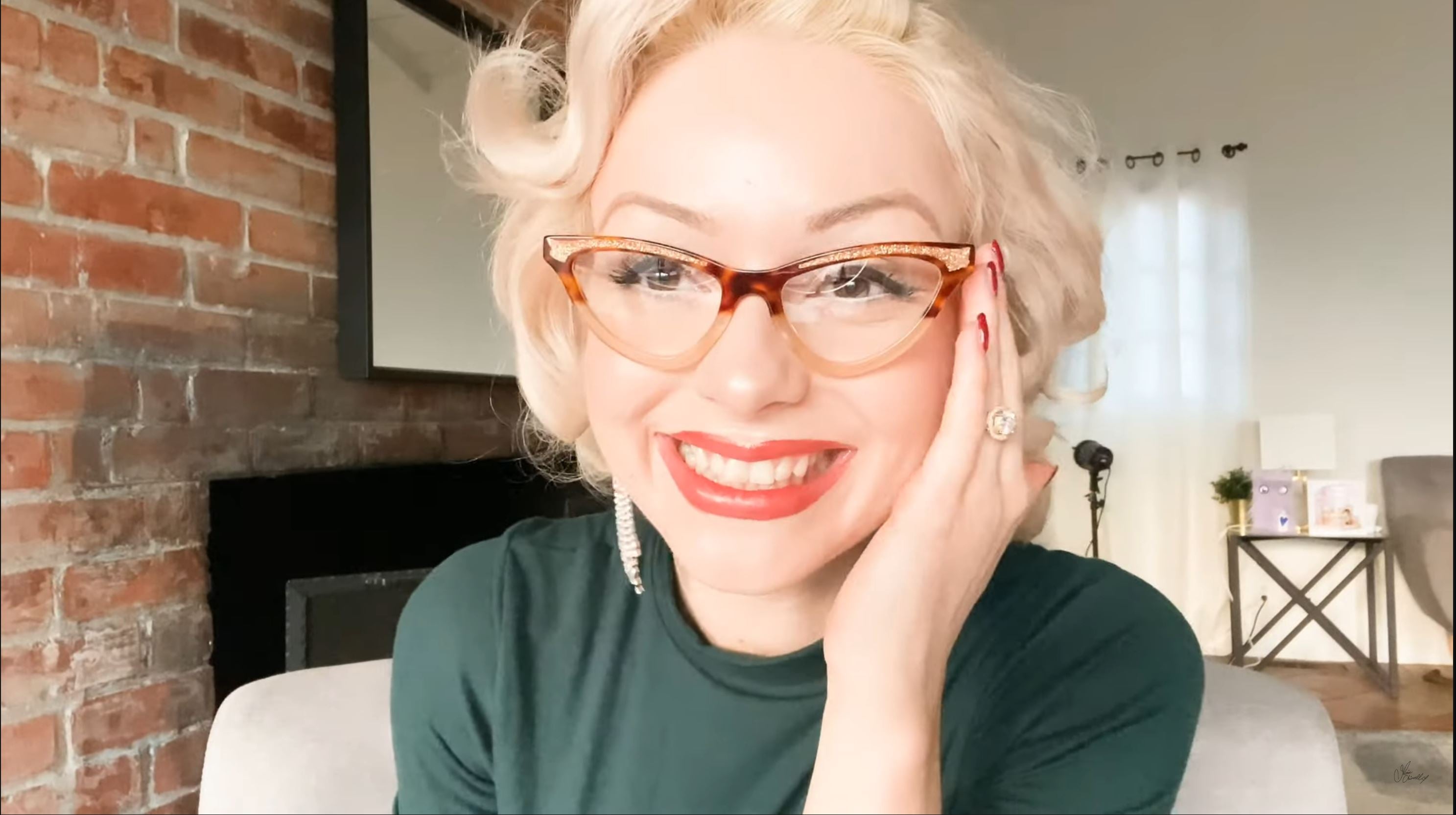 Jasmin Chiswell with JOIUSS™ cat eye glasses smiling in a room with a brick wall and modern decor.